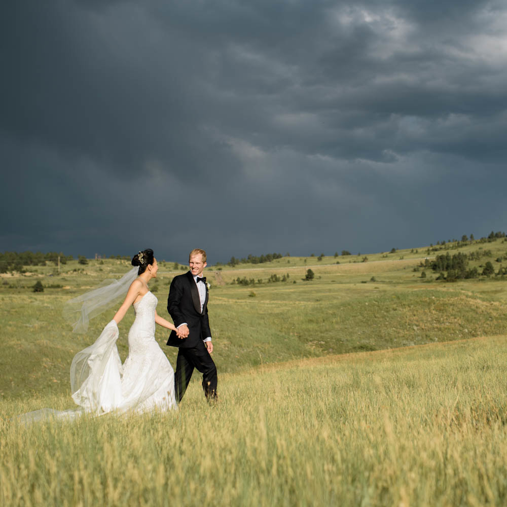 Bride and groom portrait, walking holding hands through field with mountain backdrop and stormy skies, boulder wedding planner, colorado wedding planner, classy formal real weddings, sweetly paired